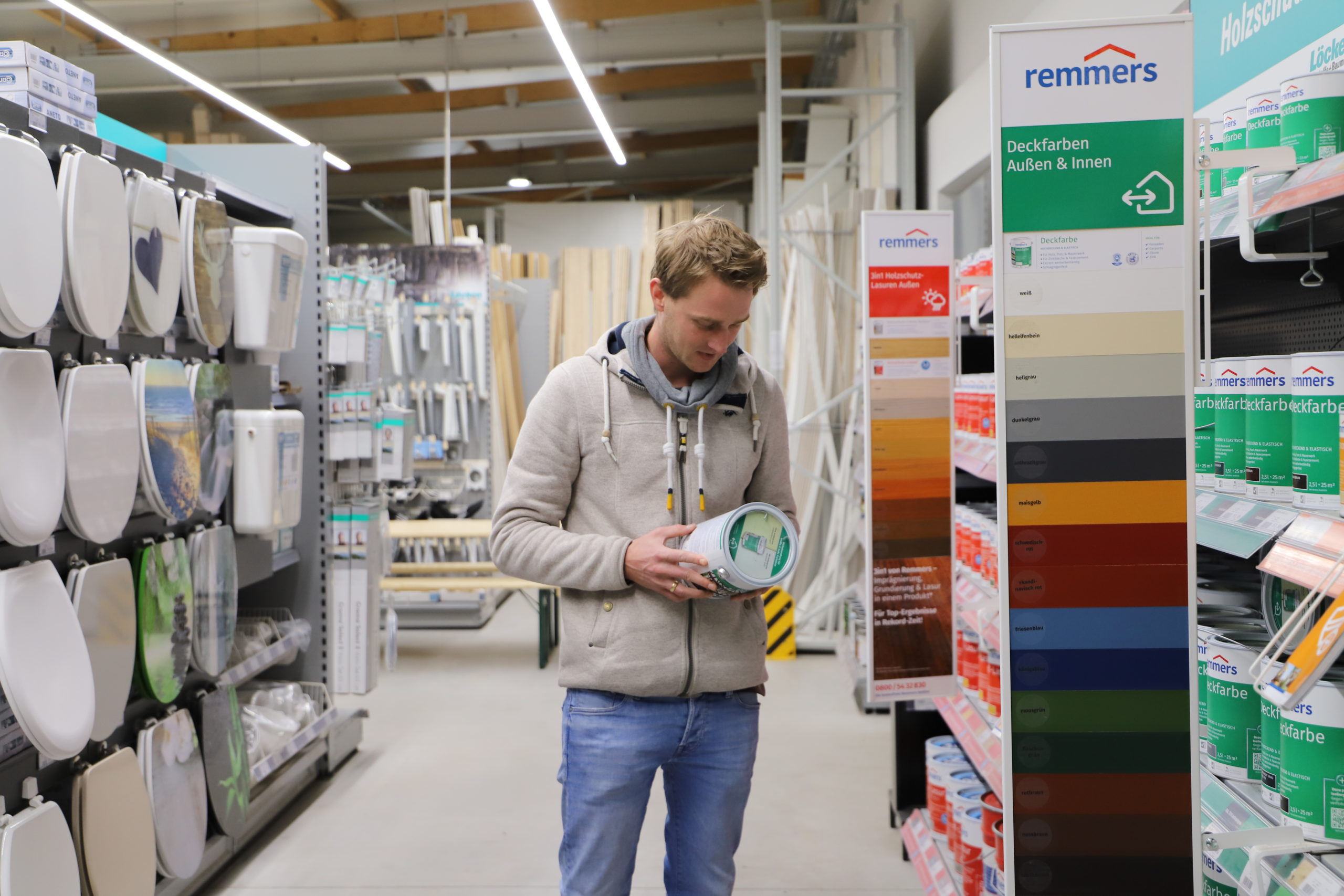 Ein Mann betrachtet eine Farbpalette und hält einen Farbeimer in einem Baumarkt, umgeben von verschiedenen Baustoffen.