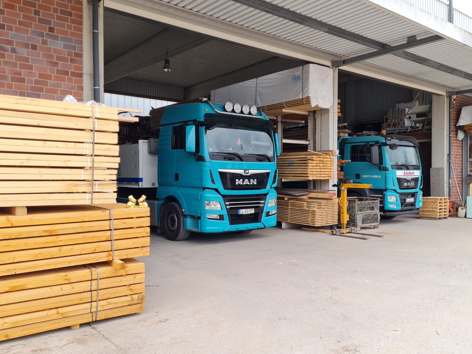 Zwei blaue LKW stehen in einem Lager, umgeben von gestapeltem Holz, bereit für den Transport von Baustoffen.