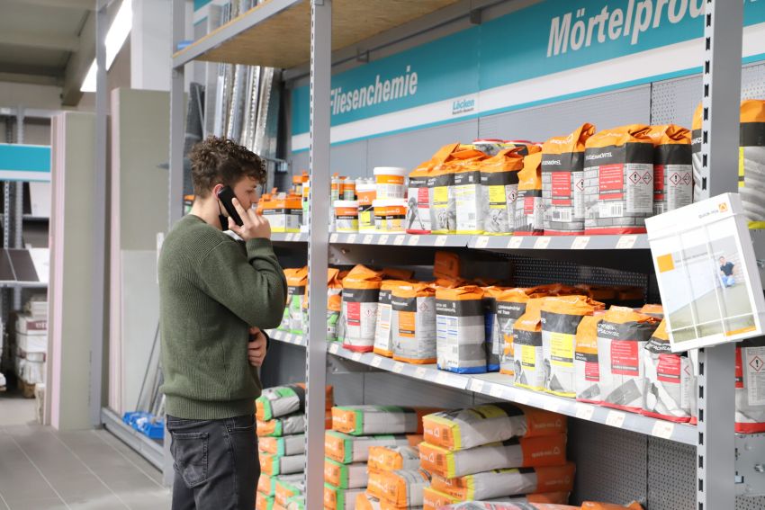 Ein Mann im Baumarkt telefoniert vor einem Regal mit Mörtelprodukten und Baustoffen, um Serviceinformationen zu erhalten.